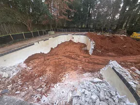 Empty pool under demolition with exposed red soil, broken concrete, construction equipment, and surrounding trees.