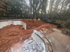 Partial demolition of a backyard pool with rubble and heavy machinery clearing red dirt near a wooded area.