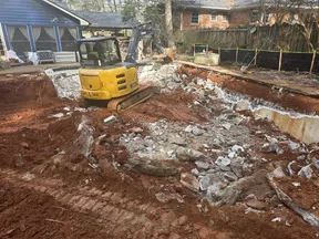 Yellow excavator demolishing a backyard pool surrounded by homes and trees with rubble and red soil