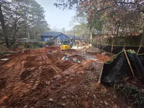 Residential backyard excavation with a yellow mini excavator clearing red soil near a blue house under overcast sky.