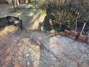 Partially constructed outdoor brick steps descending into a dirt area with surrounding bushes and a silt fence.