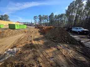 Construction site with dirt piles, tire tracks, green dumpsters, trucks, and trees under a blue sky.