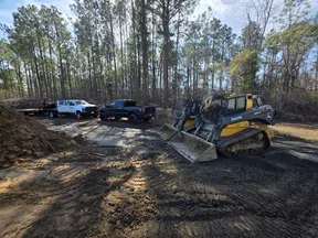 Construction site with two John Deere skid steers and two pickup trucks near a forested area under clear sky.