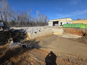 Construction site excavation with exposed foundation, rubble pile, and industrial building under clear blue sky.