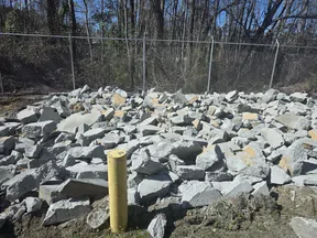 Pile of broken concrete rocks behind a yellow post with a chain link fence and trees in the background.