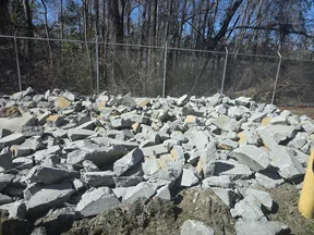 Pile of broken concrete rubble near chain-link fence with bare trees on a clear day.