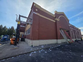 Red brick building under renovation with ladders, construction equipment, and debris on the ground under a blue sky.
