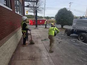 Construction workers wearing safety gear cleaning debris from a sidewalk near a building with a red dumpster and lift truck in background