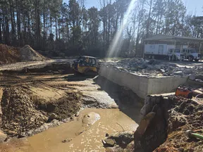 Construction site with excavator digging near concrete foundation and muddy water under sunlight with forest background