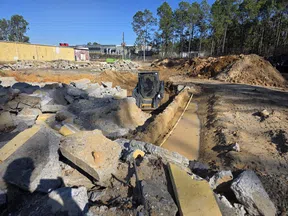 Construction site with excavator digging trench surrounded by broken concrete and dirt piles under blue sky.