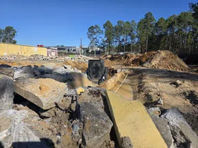 Construction site with excavator digging soil trench amid broken concrete slabs and dirt piles under clear blue sky.