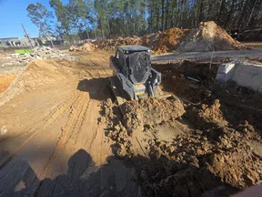 Compact bulldozer moving soil at a construction site surrounded by dirt mounds and forest trees.