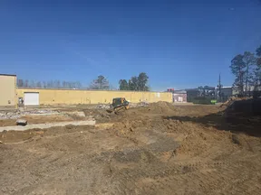 Construction site with dirt piles and a bulldozer working near a yellow building under clear blue sky.