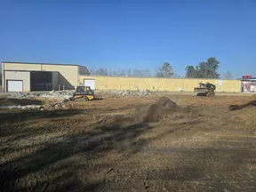 Construction site with heavy machinery working on dirt ground near a large yellow industrial building under clear blue sky.