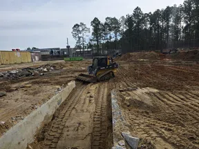 Bulldozer leveling dirt on a construction site with tire tracks and equipment in a cleared area near trees.