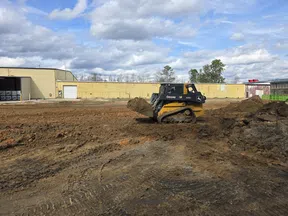 John Deere skid steer loader moving dirt at a construction site with industrial buildings and partly cloudy sky.