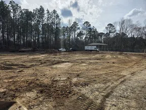 Construction site with cleared dirt ground, tire tracks, trucks, and a forested background under a cloudy sky