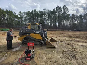 Man pressure washing a yellow John Deere skid steer loader at a muddy construction site with trees in background.