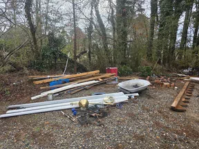 Construction materials including wood beams, metal sheets, bricks, and a wheelbarrow on a gravel lot surrounded by trees.