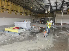 Construction worker cutting concrete floor inside large unfinished warehouse with insulation and drywall materials.