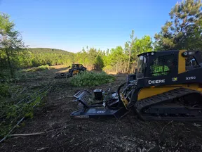 John Deere skid steer loaders clearing brush in a forested area under a clear blue sky.