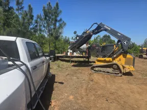 Yellow John Deere 335 compact track loader operating near a white pickup truck in a dirt clearing with trees in the background.
