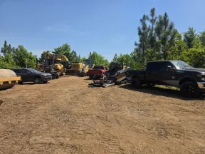 Construction vehicles and trucks parked on a dirt lot surrounded by green trees under a clear blue sky