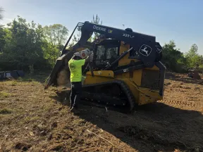 Worker operating John Deere 333 P skid steer loader clearing land on a sunny day surrounded by trees.