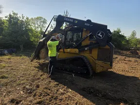 Construction worker operating a John Deere 333 LP compact track loader on a dirt site surrounded by trees under clear sky.