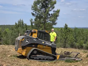 Man in neon yellow shirt standing next to a John Deere 333G compact track loader with forest background.