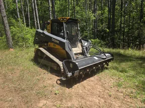 John Deere compact track loader clearing land in a forested area on a sunny day.