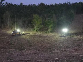 Two excavators with bright lights clearing land near a forest edge at dusk