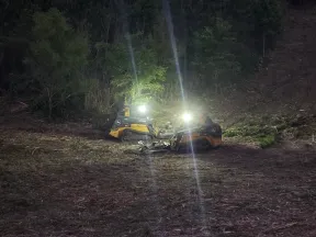 Two illuminated skid steer loaders working at night clearing a forest area with trees in the background.