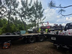 Construction workers and equipment on a trailer in a wooded outdoor worksite under a cloudy sky