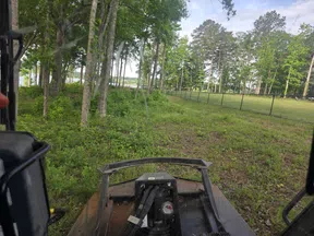 View from lawn mower cutting grass near a fence and trees under a partly cloudy sky.