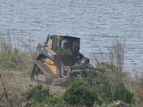 John Deere tracked skid steer clearing vegetation near a water body on rugged terrain.