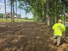 Man in neon shirt inspecting cleared land near trees with a house and pond in the background on a sunny day.