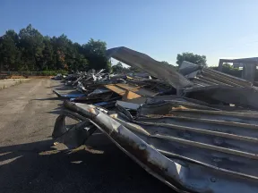 Piles of twisted and dented metal sheets lying in an outdoor lot with trees and clear blue sky.
