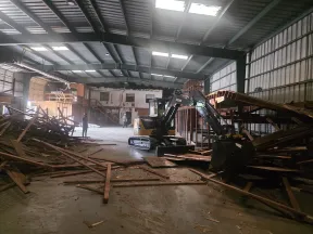 Indoor construction site with excavator clearing wooden debris inside a large warehouse with workers present