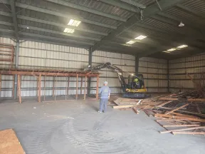 Worker operating excavator inside warehouse clearing wooden debris from metal shelving demolition.
