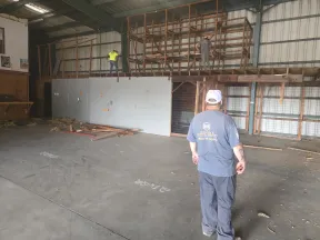 Three construction workers renovating a large indoor warehouse with wooden scaffolding and concrete floor under metal roof.