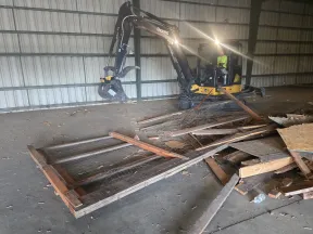 Mini excavator inside a warehouse demolishing wooden debris and fencing materials on concrete floor.