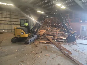 John Deere excavator inside warehouse clearing a large pile of wooden debris and lumber pieces scattered on the floor.