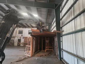 John Deere excavator demolishes wooden structure inside large metal warehouse building with debris on floor.