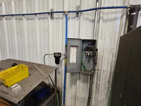 Industrial workspace with metal table, yellow toolbox, open electrical panel, and blue piping mounted on wall.