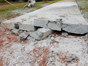 Cracked and broken concrete sidewalk with rubble and construction equipment nearby during repair work.