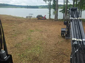 View from machinery at cleared lakeside land with trees, boat dock, and equipment under cloudy sky.