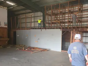 Three workers in a warehouse are dismantling a large wooden shelf structure with tools and debris on the floor.