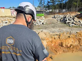 Construction worker in safety helmet operating hose near muddy water pit on demolition site with rubble piles.