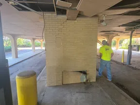 Cracked brick pillar with broken ceiling panels and worker in bright shirt in a parking garage repair scene.